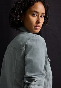 Jasnoszara kurta z korduroyu z kołnierzykiem i guzikami. Pokazana z boku, z teksturowanym materiałem i luźnym krojem.