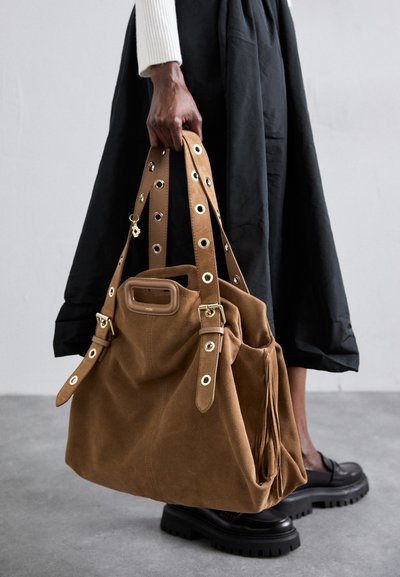 Bolso tote de ante marrón con herrajes dorados, presenta correas con tachuelas y un bolsillo frontal. Acompañado de una falda negra y zapatos gruesos.