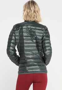 Osoba s krátkými blond vlasy, oblečená v tmavě zelené prošívané bundě a červených kalhotách, viděná zezadu na bílém pozadí.