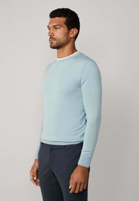 Muž v světle modrém svetru s dlouhým rukávem a tmavě modrých kalhotách, stojící bokem na jednobarevném světlém pozadí.