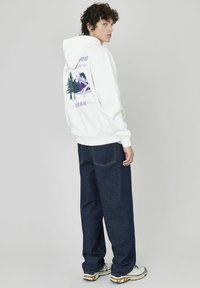 Un hoodie blanc avec un graphique de montagne et d'arbre en violet et vert au dos, associé à un jean large bleu foncé et des baskets.