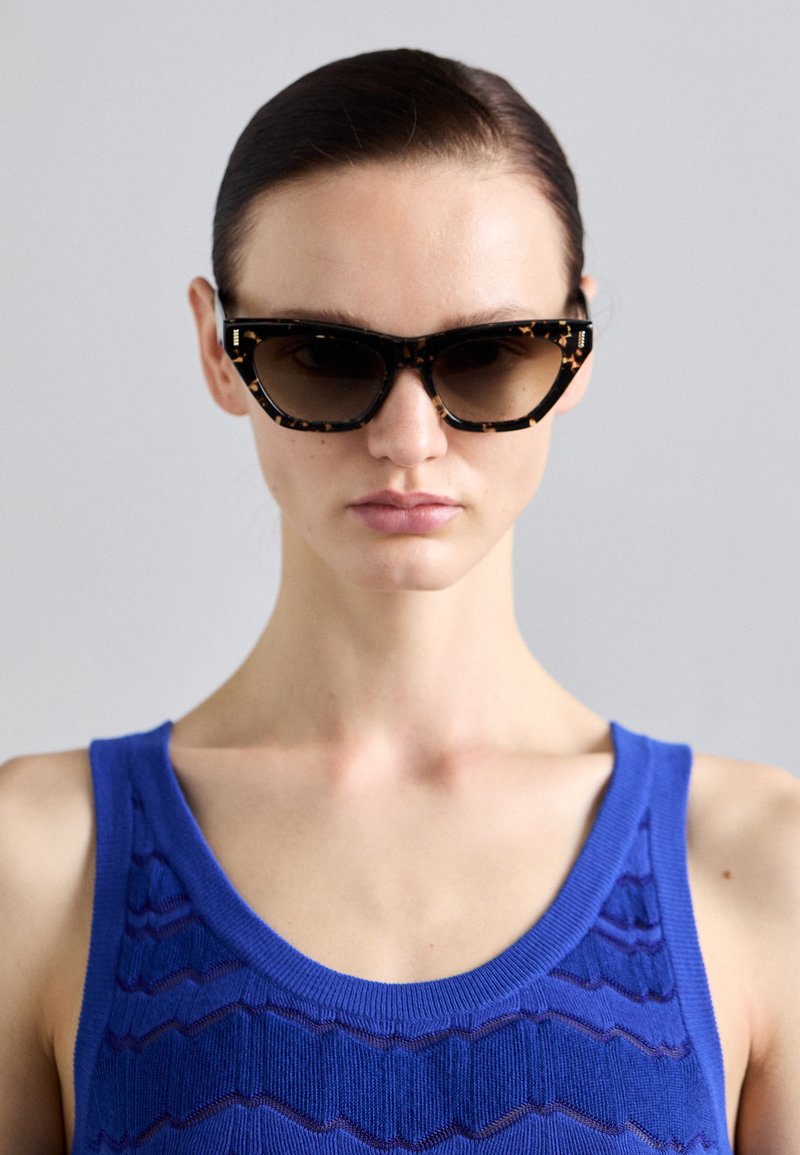 Occhiali da sole tartarugati con lenti scure, caratterizzati da una forma a occhio di gatto audace e montature spesse. Il modello indossa un top senza maniche blu e texturizzato.