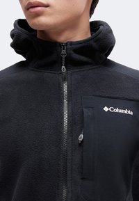 Κοντινό πλάνο ενός ατόμου που φοράει μαύρο fleece μπουφάν Columbia με ψηλό γιακά και μπροστινό φερμουάρ, εμφανίζοντας το λογότυπο της μάρκας στην τσέπη στο στήθος.