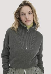 Grauer Wollpullover mit hohem Kragen und halbem Reißverschluss, der im oberen Bereich eine gerippte Textur aufweist und eine lockere Passform hat. Über einem grünen Rollkragenpullover getragen.