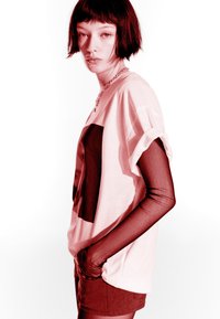 Mladý človek s krátkymi čiernymi vlasmi, oblečený v bielom oversized tričku cez priesvitný dlhý rukáv a šortkách, stojí v bočnom profile.