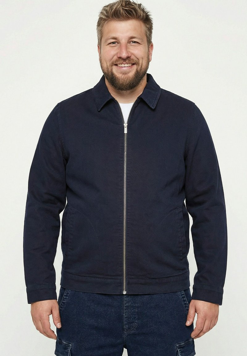 Lächelnder Mann mit kurzen blonden Haaren und Bart, der eine dunkelblaue Reißverschlussjacke und dunkle Jeanshosen vor einem schlichten weißen Hintergrund trägt.