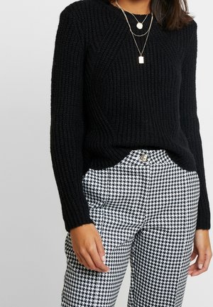 Femme portant un pull côtelé noir, des colliers en or superposés et un pantalon taille haute à motif pied-de-poule noir et blanc avec poches avant et fermeture à boutons.