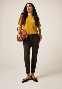 Haut tricot jaune à manches courtes, pantalon marron ajusté, ballerines noires à bouts pointus avec des accents marron, tenant un sac à main marron. Design texturé simple.