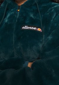 Zielona kurtka z sztucznego futra na zamek z czarnym haftowanym logo "ellesse". Posiada kaptur oraz boczne kieszenie. Miękka faktura z błyszczącym wykończeniem.