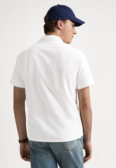 Balta īsroku krekls ar apkakli, valkāts kopā ar zilām džinsu biksēm. Uz galvas ir tumši zila cepure. Audums izskatās mīksts ar gludu tekstūru.