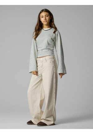 Jeune femme debout portant un haut à manches longues rayé noir et blanc, un pantalon large beige taille haute et des chaussures plates marron foncé sur fond gris.