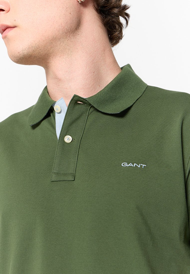 Polo shirt vert en tissu texturé, avec un col bleu clair, deux boutons et un logo blanc brodé sur la poitrine.
