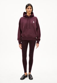 Bordeauxkleurige hoodie met een voorzak en een witte lintlogo, gecombineerd met zwart en paarse zebra-gestreepte legging en zwarte loafers.