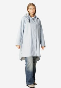 Jasnoniebieski, wodoodporny płaszcz przeciwdeszczowy z kapturem, zatrzaskami i bocznymi kieszeniami. Noszony na wyblakłych niebieskich dżinsach i beżowych butach.