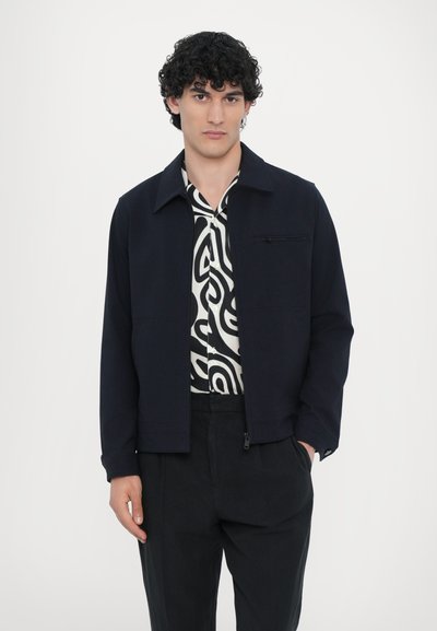 Modra jakna z ovratnikom in zadrgo, v kombinaciji z srajco v črno-belem vzorcu. Ima sprednji žep in ohlapen kroj.