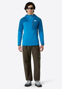Giacca blu con zip, in tessuto testurizzato e dettagli arancioni, abbinata a pantaloni marroni e sneakers grigie. Indossa occhiali da sole.