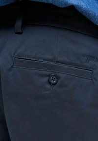 Κοντινό πλάνο σε σκούρη μπλε τσέπη παντελονιού με κούμπωμα κουμπιού και διακριτικό κεντημένο λογότυπο κοντά στη μέση κάτω από ένα μπλε πουκάμισο.