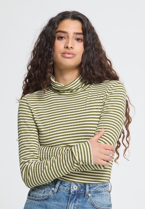 Femme aux longs cheveux bouclés portant un col roulé rayé vert et blanc et un jean bleu, debout les bras croisés devant un fond uni.