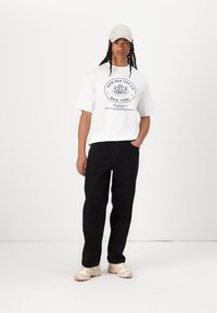 Bijela grafička majica kratkih rukava s plavim logom, crne hlače i bež tenisice. Odjevna kombinacija ima ležeran kroj i neutralnu paletu boja.