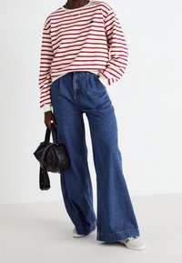 Camicia a maniche lunghe a righe rossa e bianca, abbinata a pantaloni in denim blu a gamba larga. Gli accessori includono una piccola borsa nera.