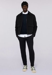 Giacca nera in misto lana con colletto, indossata sopra un maglione blu navy e una camicia bianca, abbinata a pantaloni neri e sneakers con dettagli verdi.