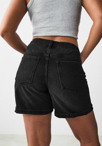 Bermudas de mezclilla negra con dobladillo enrollado, que cuentan con dos bolsillos traseros, cintura alta y una textura suave. Diseño ligero y casual.