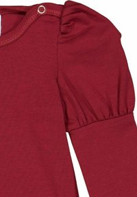 T-shirt rouge à manches longues avec épaules bouffantes, tissu texturé et fermeture à pression au niveau du cou. Texture lisse avec des détails froncés.