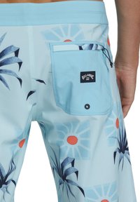 Costumi da bagno blu con un motivo floreale caratterizzato da accenti arancioni e blu scuro. Include una tasca sul retro con un'etichetta del logo.