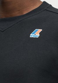 Κοντινό πλάνο ενός ατόμου που φοράει μαύρο μπλουζάκι με στρογγυλή λαιμόκοψη και ένα μικρό μπλε και πορτοκαλί λογότυπο K-Way στο πάνω μέρος του στήθους.