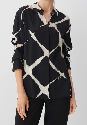 Czarna bluzka zapinana na guziki z dużym nieregularnym białym wzorem krzyżowym, noszona z podwiniętymi rękawami do łokci i zestawiona z czarnymi spodniami.