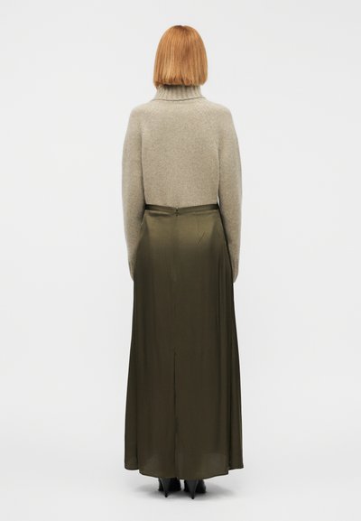 Πλεκτό μπεζ πουλόβερ με ζιβάγκο σε συνδυασμό με μακριά φούστα σε ελιά χρώμα, με σχισμή στο πίσω μέρος, λεία υφή και εφαρμοστή μέση. Φαίνονται μαύρα τακούνια.