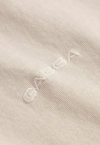 Helles beiges Gewebe mit einer strukturierten Webart, das hervorgehobene weiße Stickerei des Wortes "GABA" in einer modernen Schriftart zeigt.