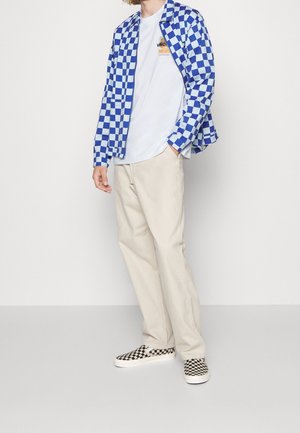 Person wearing blue and white checkered jacket, white graphic t-shirt, beige pants, and black and white checkered slip-on shoes.