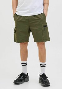 Pantaloni scurți cargo de culoare verde oliv, cu multiple buzunare, fermoare și un talie plată. Purtați cu încălțăminte sport negre și șosete albe cu dungi.