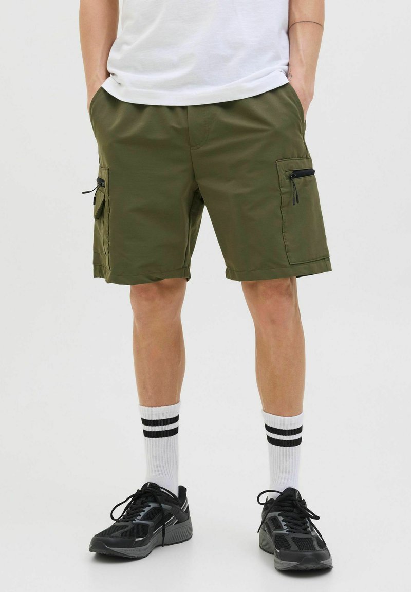 Pantaloni scurți cargo de culoare verde oliv, cu multiple buzunare, fermoare și un talie plată. Purtați cu încălțăminte sport negre și șosete albe cu dungi.