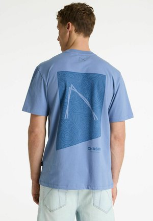 Mees kannab helesinist T-särki, millel on topograafilise kaardi kujundus ja "CHASIN" logo taga, koos heledate teksadega.