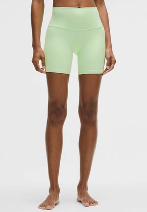 Vrouw draagt lichtgroene, hooggetailleerde fietsbroek tot halverwege de dij, staand op blote voeten tegen een effen witte achtergrond.