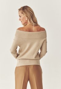 Beżowy sweter ze szwami na ramionach z prążkowanym dekoltem i mankietami. W zestawie z beżowymi spodniami o podwyższonym stanie z subtelnymi tylnymi kieszeniami.