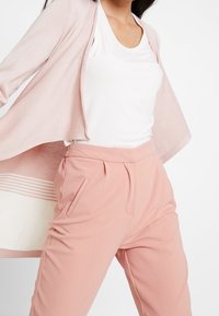 Žena v světle růžovém cardigan, bílém topu bez rukávů a růžových kalhotách s vysokým pasem na bílém pozadí.