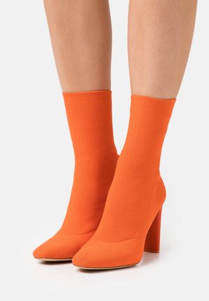 Bottes à chevilles de style chaussette orangées avec un bout pointu et un haut talon carré. Fabriquées en tissu extensible, elles présentent une texture lisse et un design sans coutures.