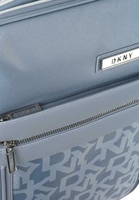 Bolso DKNY gris con una superficie texturizada y un logotipo prominente. Incluye un bolsillo con cremallera y un patrón en relieve.