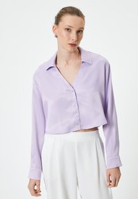 Lilla cropped bluse med lange ærmer, knaplukning, glat satin tekstur og klassisk krave. Kombineret med hvide bukser.