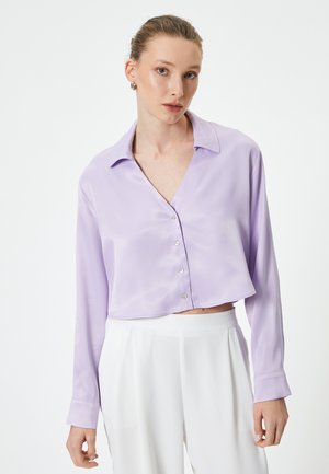 Blusa corta lilla con maniche lunghe, chiusura con bottoni, texture in satin morbido e colletto classico. Abbinata a pantaloni bianchi.