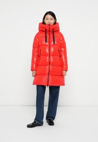Rote Puffjacke mit glänzender Oberfläche, übergroßer Kapuze, schwarzen Kordeln und Reißverschluss-Taschen. Kombiniert mit lockeren blauen Jeans.