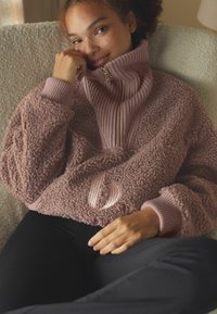 Flísová mikina v barvě prachové růže s vysokým límcem, žebrovanými detaily a měkkou texturou. Má zipové zapínání a vyšívané logo.