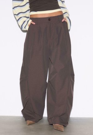 Osoba ubrana w szerokie, brązowe spodnie z zakładkami z przodu, ręce w kieszeniach, w połączeniu z brązowymi, spiczastymi butami oraz krótkim paskowym topem.