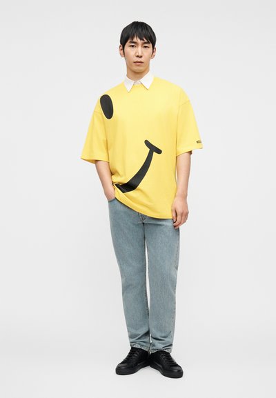 Κίτρινη υπερμεγέθη μπλούζα με μεγάλο σχέδιο ενός χαμογελαστού προσώπου και μαύρες λεπτομέρειες, συνδυασμένη με ανοιχτό μπλε τζιν παντελόνι και μαύρα αθλητικά παπούτσια.