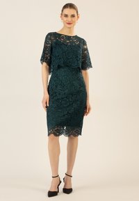 Teal Spitzenkleid mit kurzen Ärmeln, das florale Muster, eine figurbetonte Silhouette und einen gewellten Saum aufweist. Kombiniert mit schwarzen High Heels.