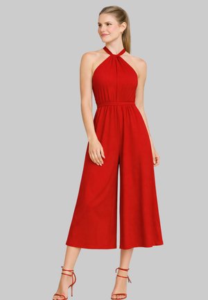 Kobieta ubrana w czerwoną bezrękawnikową kombinezon-halterskę z szerokimi, skróconymi nogawkami, oraz czerwone wysokie obcasy z paskiem wokół kostki, stojąca na szarym tle.