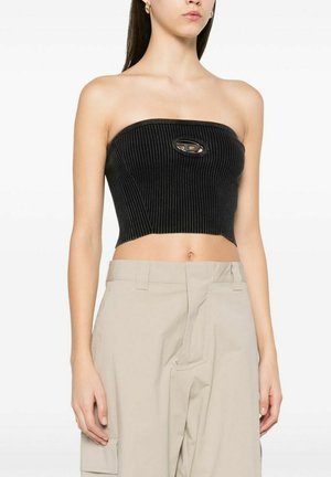 Donna che indossa un crop top nero a coste senza spalline con un piccolo taglio e pantaloni beige con vita alta e tasche laterali, in piedi davanti a uno sfondo bianco.